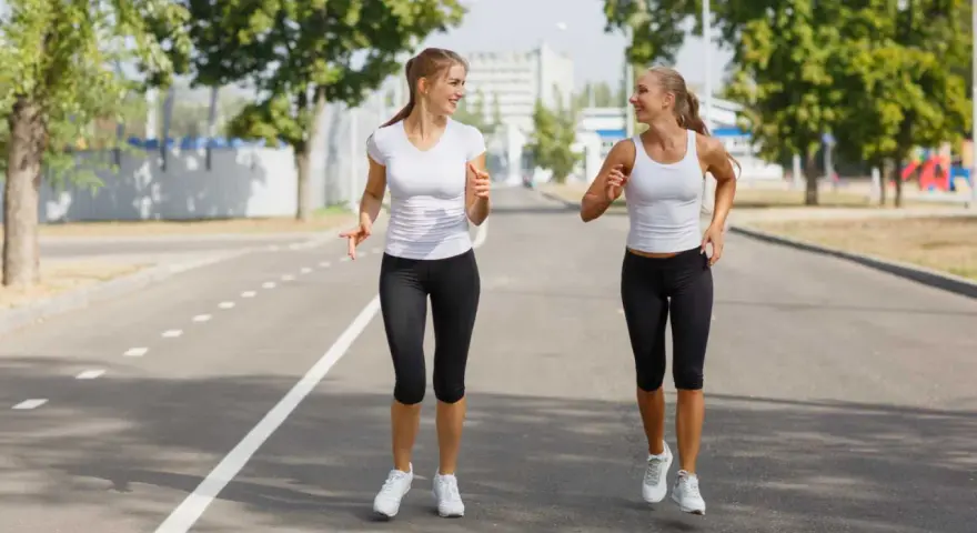 Due donne sorridono mentre praticano diversi tipi di camminata sportiva su una strada asfaltata, con alberi e edifici sullo sfondo.