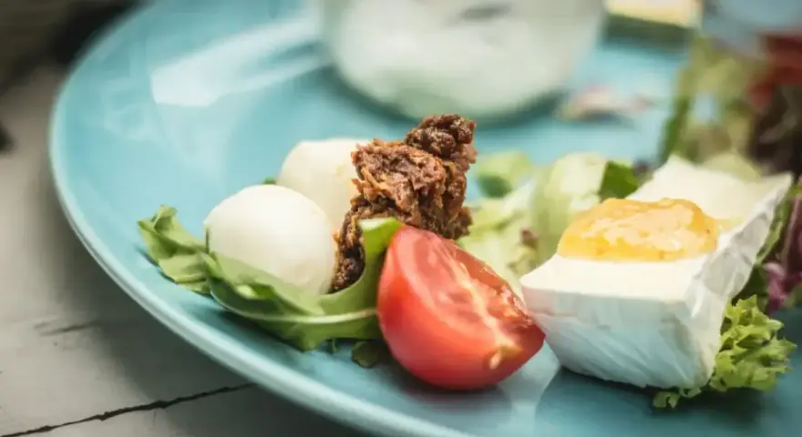 Pyszne keto śniadanie: ser brie z dżemem, kulki mozzarelli, pomidor i rukola na błękitnym talerzu.