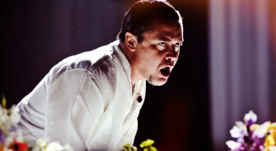 Mike Patton heute, mit weit aufgerissenem Mund und intensivem Blick, trägt ein weißes Hemd. Blumen im Vordergrund.