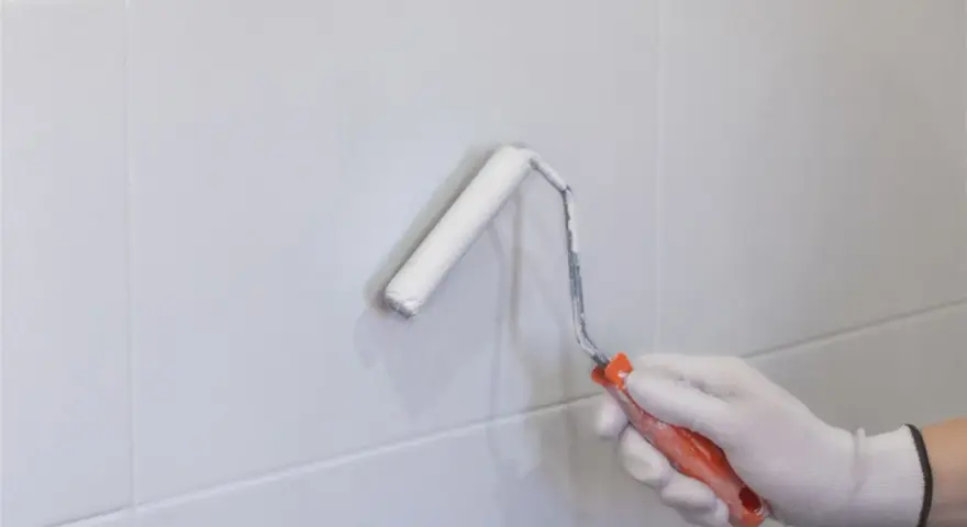 Mano enguantada pintando con rodillo blanco sobre azulejos grises. Renovando el aspecto de los tubos calefacción.