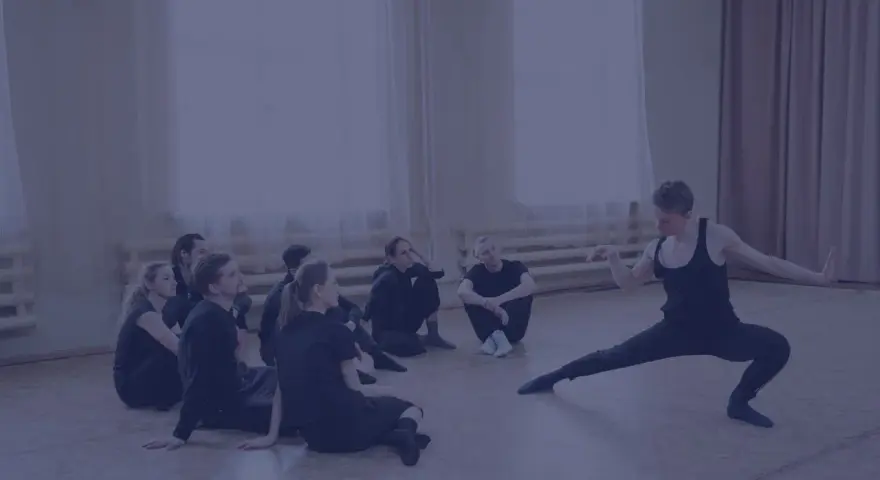 Choreograf demonstruje ruchy grupie tancerzy siedzących na podłodze.