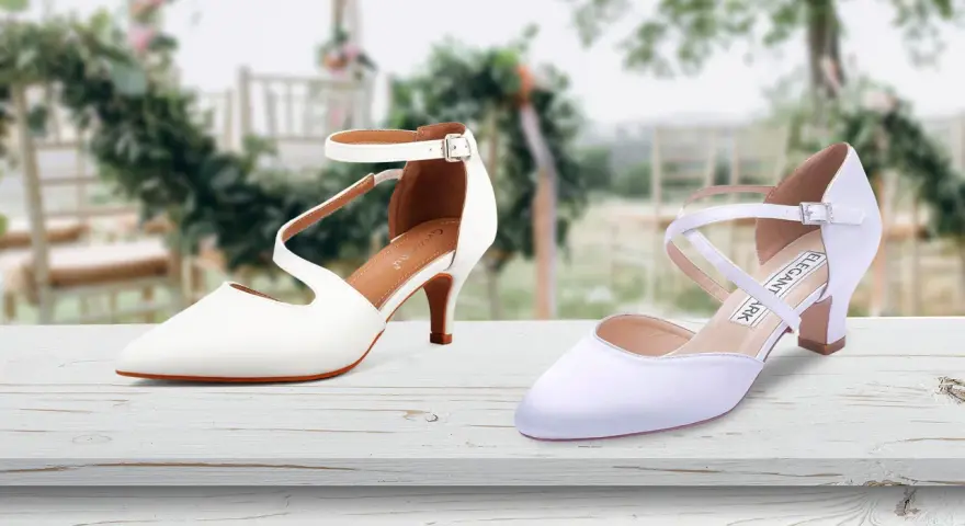 Due eleganti scarpe da sposa bianche, una con cinturino alla caviglia, l'altra con incroci sul collo del piede, su un tavolo di legno con sfondo sfocato di un matrimonio.