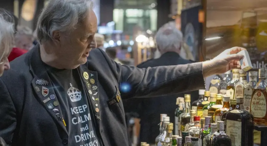Mann mit "Keep Calm and Drink Whisky"-Shirt und vielen Ansteckern auf einer Whiskey Messe, der eine kleine Box hält.