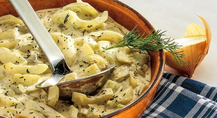 Cremiges Schmorgurken Rezept mit Dill in einer Tonschale, daneben eine Zwiebel und ein Löffel.
