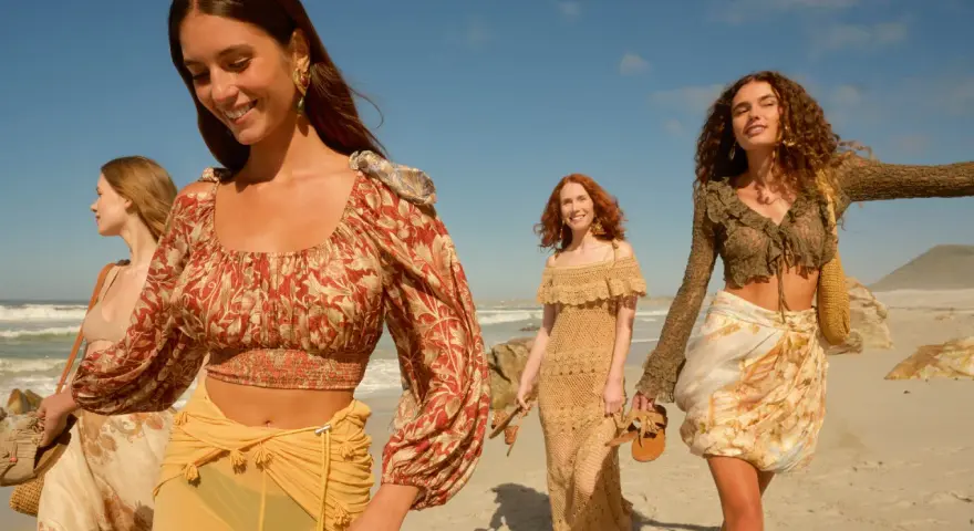 Quattro donne con abiti in stile boho passeggiano su una spiaggia sabbiosa con il mare sullo sfondo.