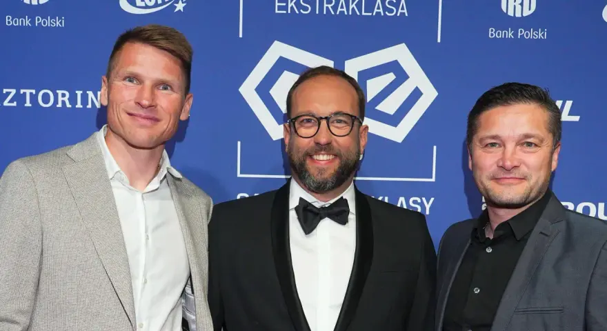 Trzech mężczyzn na tle PKO Bank Polski Ekstraklasa. Jeden z nich, w garniturze, uśmiecha się do obiektywu, być może jako jeden z przyszłych królowie strzelców ekstraklasy.