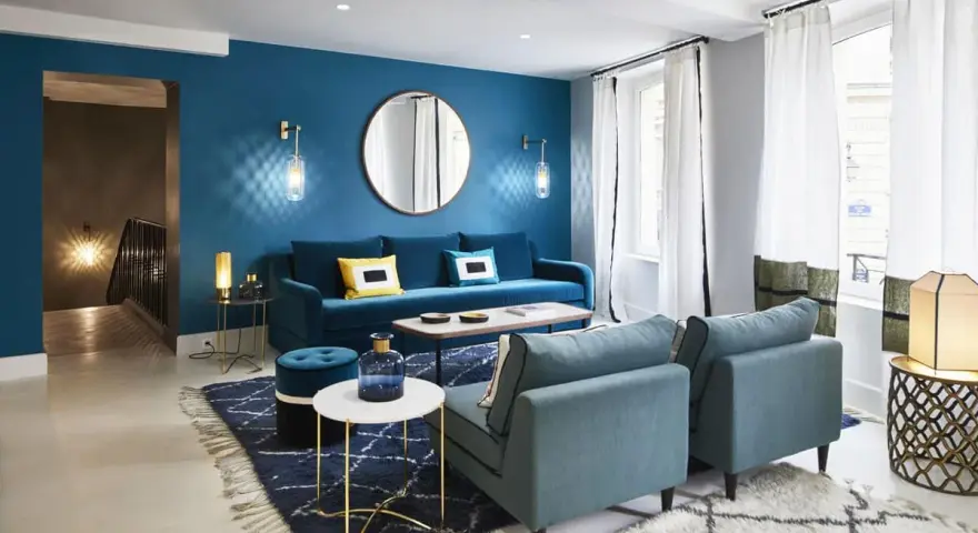 Salon moderne avec mur bleu canard, canapé velours, fauteuils et tapis assortis. Le jaune vif des coussins dynamise l'ensemble.