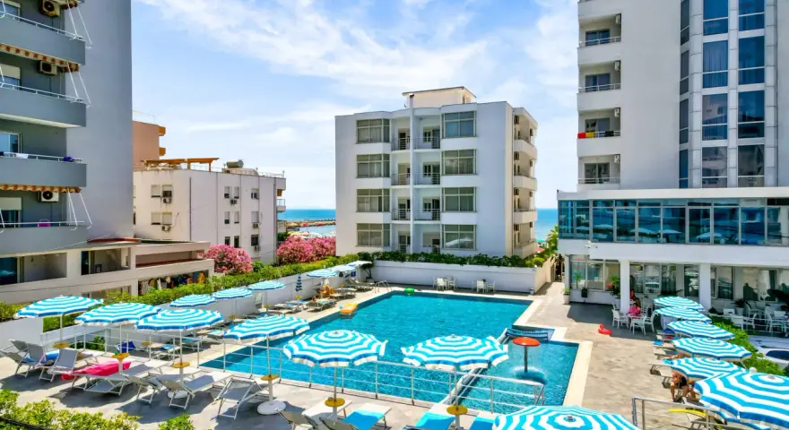 Albania - wczasy samolotem all inclusive. Basen hotelowy z leżakami i parasolami, w tle budynki i morze. Idealne miejsce na relaks.