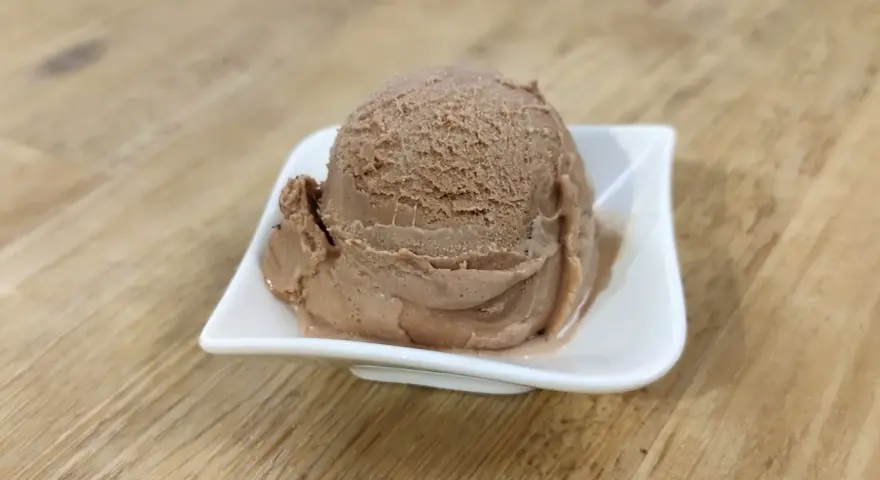 Ein Löffel cremiges Nougat Eis in einer weißen Schale auf einem Holztisch.