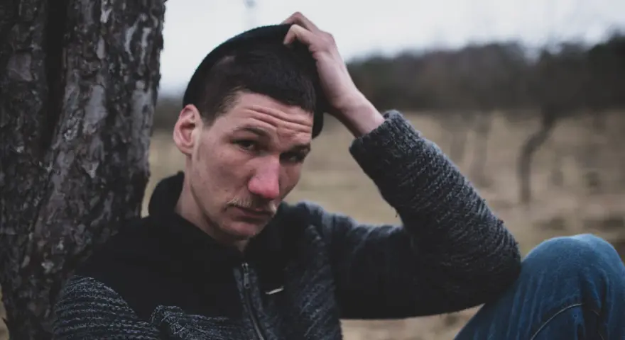 Quebonafide w czapce i swetrze, oparty o drzewo. W tle jesienny krajobraz. Tekst "żadnych zmartwień" pasuje do nastroju.