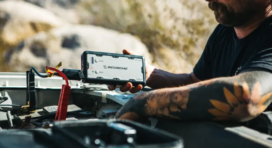 Man with tattoos demonstrates how to charge a jump starter by connecting it to a car battery in a desert landscape.