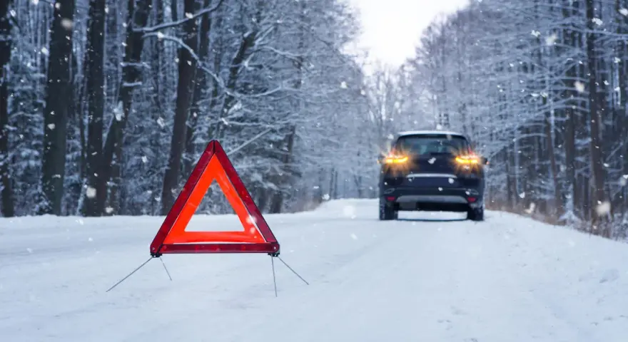 Warnblinkleuchten und Warndreieck auf verschneiter Straße. Ein Auto steht im Hintergrund. Ein wichtiger Hinweis für den auto wintercheck.