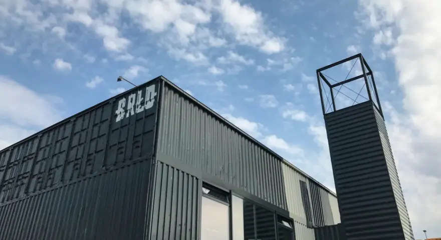Moderne Containerarchitektur am **brlo gleisdreieck** mit Schriftzug "BALD" und einem markanten Turm unter blauem Himmel.