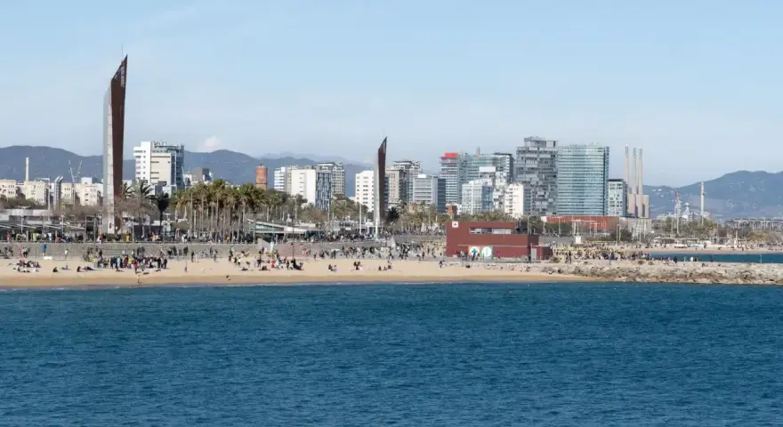 Ludzie odpoczywają na plaży, a w tle widać nowoczesne budynki Barcelony. To miasto leży nad Morzem Śródziemnym.
