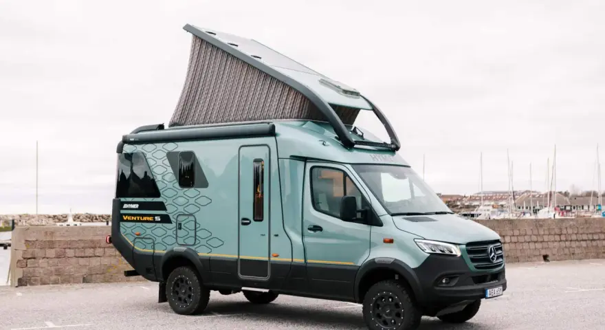 En grön Hymer Venture S husbil med uppfällt tak står parkerad vid en hamn.