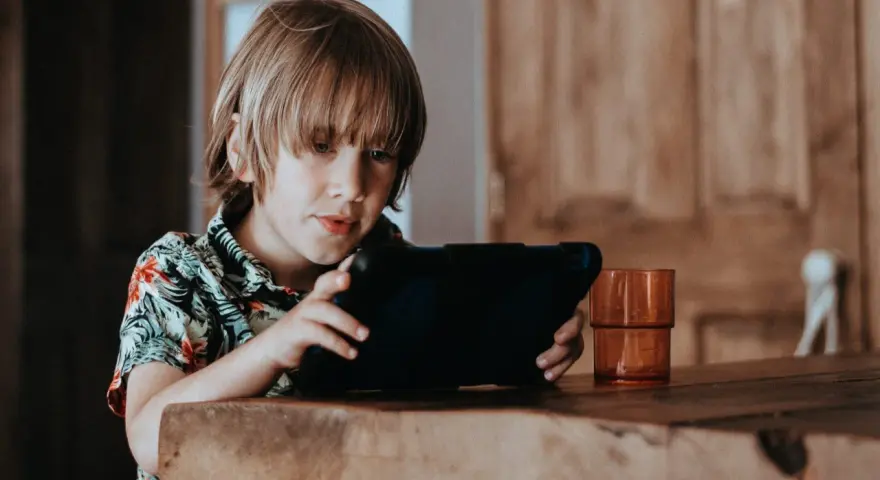Chłopiec w hawajskiej koszuli ogląda krótkie filmy na tablecie, siedząc przy drewnianym stole.