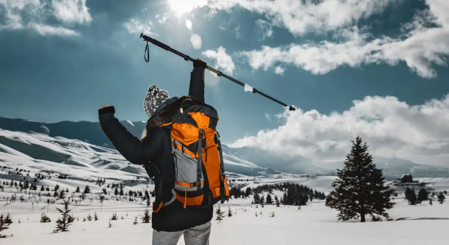 Osoba z plecakiem wznosi kijek, przygotowując się do marszu. Dowiedz się, jak prawidłowo założyć buciki na kijki nordic walking, by cieszyć się zimową wędrówką.