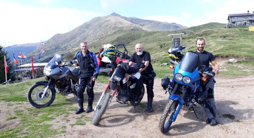 Tre motociclisti con moto da enduro posano in cima al passo San Lucio, con montagne sullo sfondo.