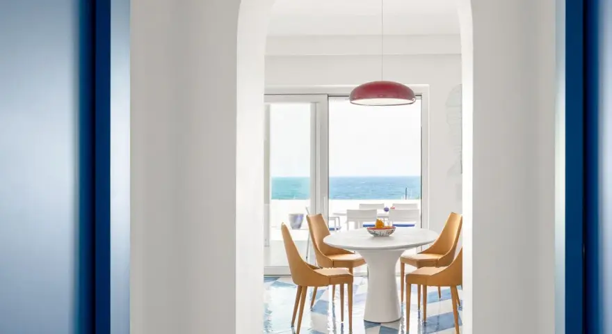 Interno di una casa stile mare con pavimento a scacchi blu e bianchi, tavolo rotondo con sedie arancioni e vista sul mare.
