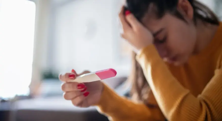 Kobieta z opuszczoną głową trzyma test ciążowy. Jej dłoń z czerwonym lakierem do paznokci zaciska się na teście. Tokofobia może wywoływać takie emocje.