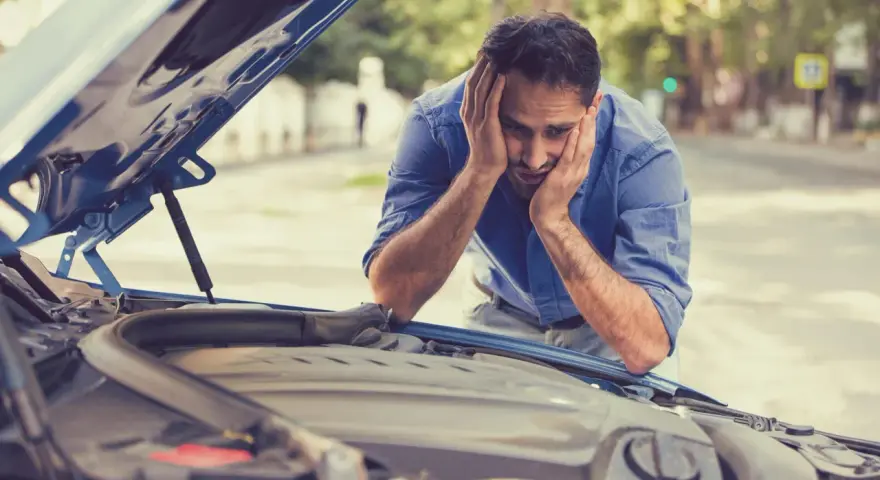 A frustrated man with hands on his head stares at his car's open hood, likely hearing a grinding noise when starting the car.
