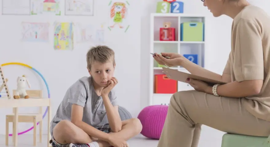 Chłopiec siedzi na poduszce, zastanawiając się, jakie pytania zadaje psycholog dziecku. W tle rysunki dzieci i kolorowe zabawki.