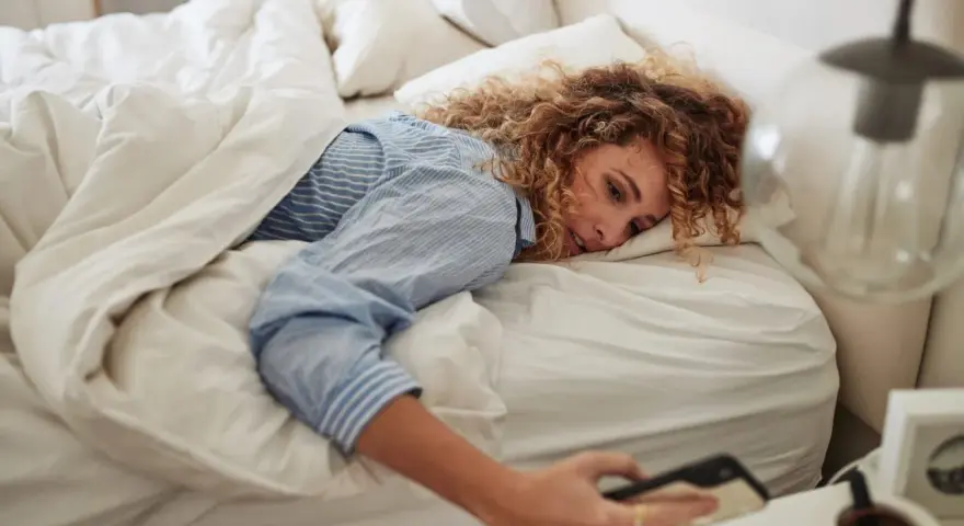 Kobieta z kręconymi włosami leży w łóżku, sięgając po telefon. Czyżby 14:44 znaczenie miało dla jej poranka?