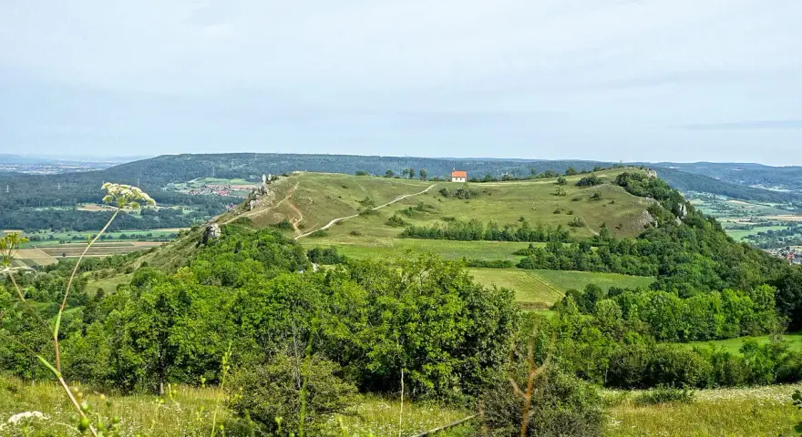 Grüne Hügel und Felder laden zum Walberla wandern ein. Ein kleiner Weg schlängelt sich zum Gipfel mit einem roten Haus.
