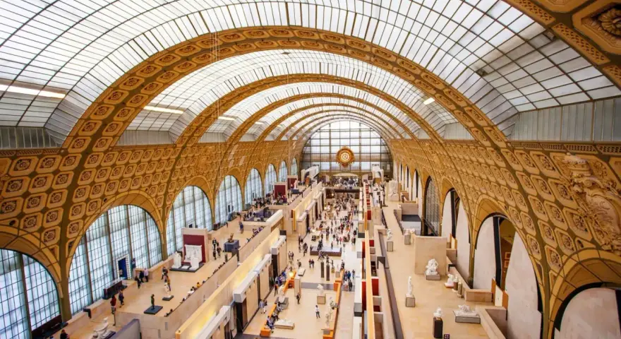 Interior del museo D'Orsay, con su impresionante techo abovedado de cristal y arcos dorados. Gente pasea entre esculturas y exposiciones.