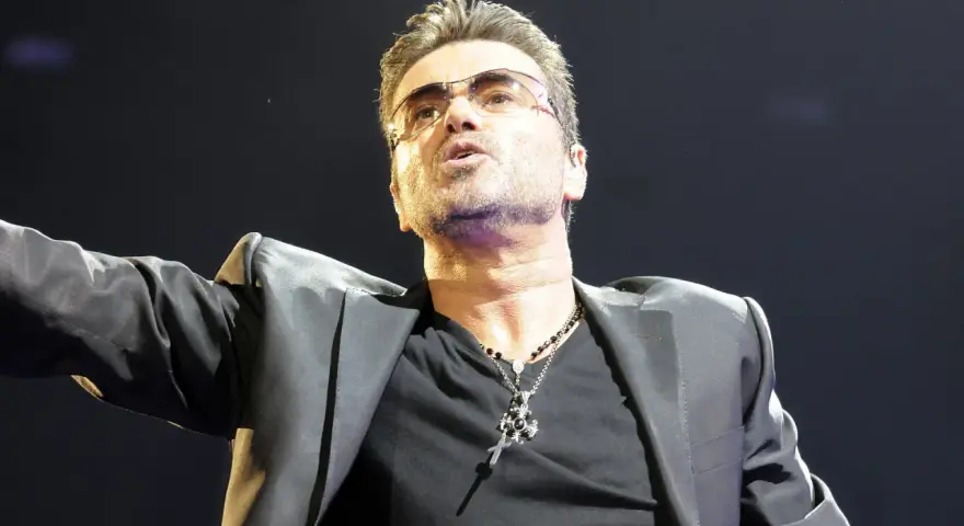 George Michael w okularach, w czarnej marynarce i z naszyjnikiem, śpiewający swoje niezapomniane utwory.