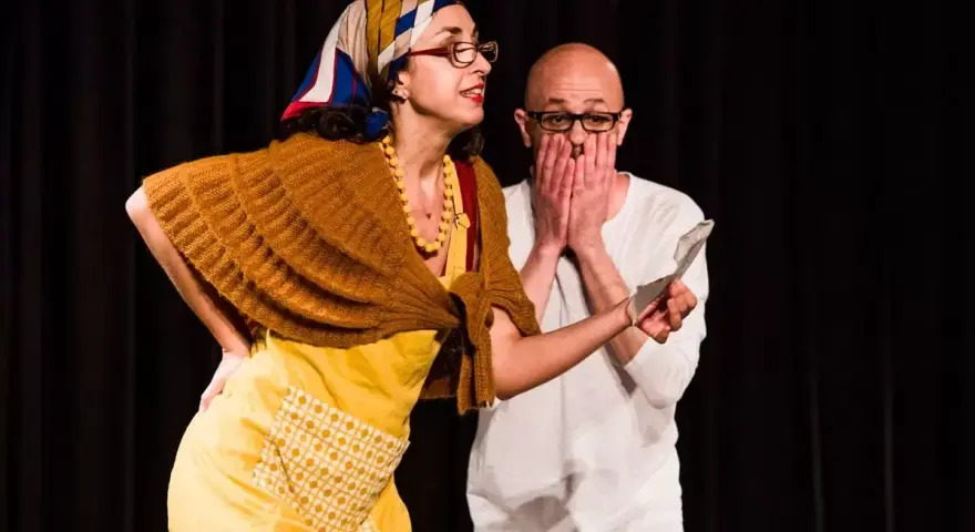 Due attori in scena, una donna con foulard e grembiule giallo, un uomo con le mani sul volto. Una scena di commedia drammatica.