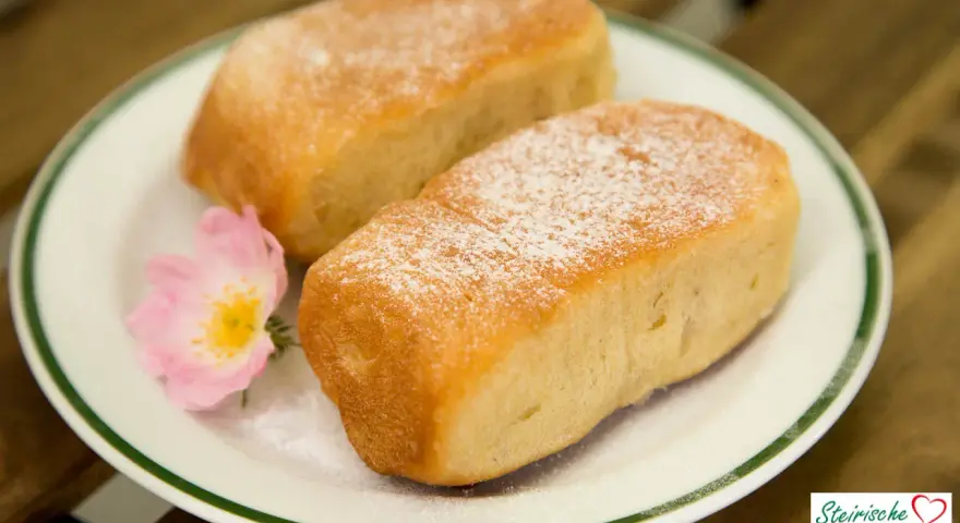 Zwei goldbraune Buchteln, bestäubt mit Puderzucker, auf einem Teller mit einer zarten rosa Blüte. Omas Rezept für Buchteln, ein Genuss!