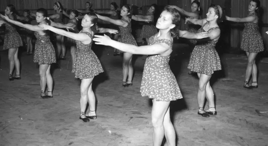 Młode tancerki w ludowych strojach ćwiczą choreografię. Ich ruchy synchronizują się z rytmem, jakby grały zespoły polskiej muzyki ludowej.
