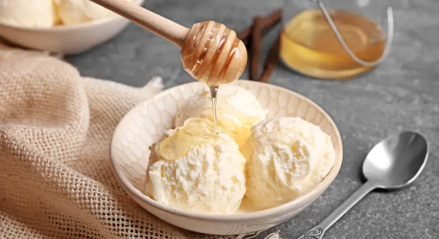 Schneeballgroße Kugeln Vanilleeis werden mit flüssigem Honig beträufelt. Ein Holzlöffel tropft goldenen Honig auf das cremige Eis.