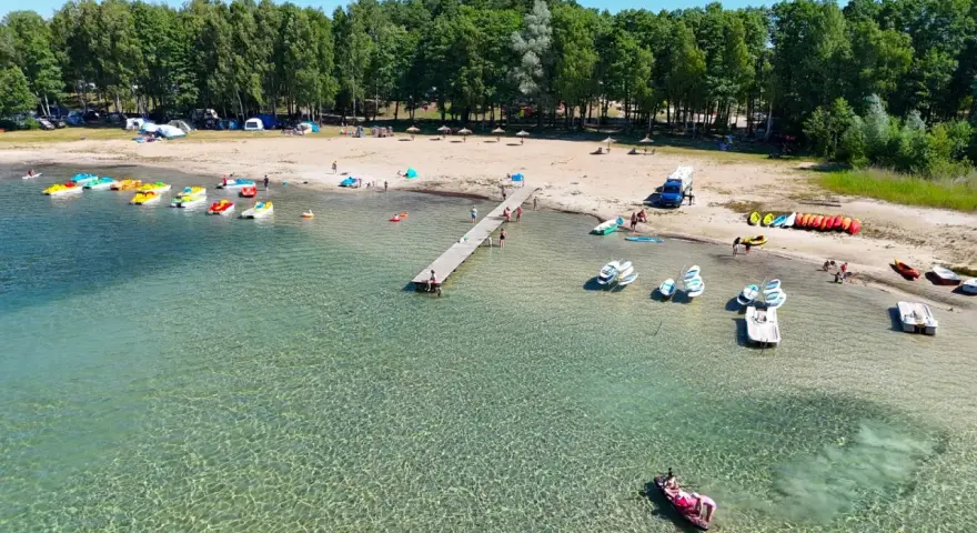 Letnia przygoda na wodzie, kolorowe rowerki wodne i kajaki czekają przy brzegu. Widok na plażę z namiotami i lasem, idealne miejsce na wypoczynek nad jeziorem Binduga 69.