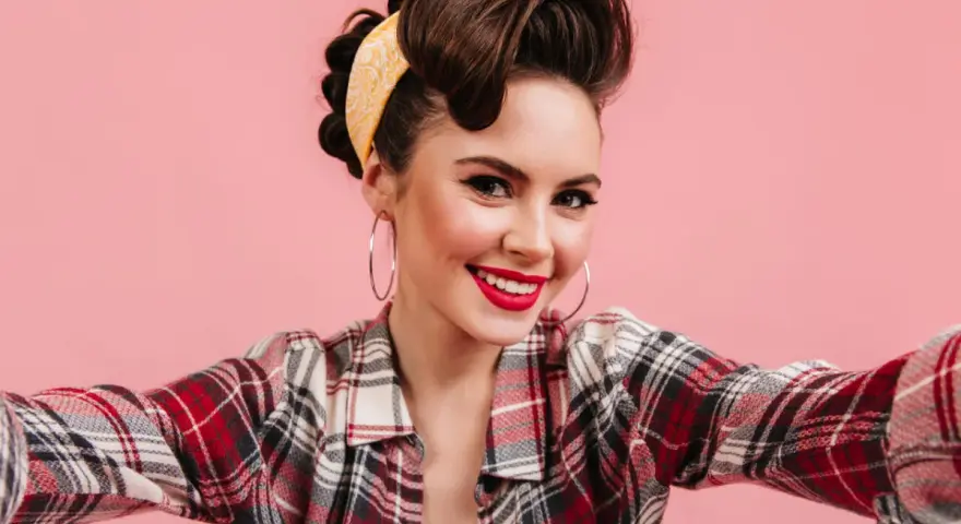 Uśmiechnięta kobieta w stylu pin-up, z burzą brązowych włosów spiętych w kok i żółtą bandaną, w kratę koszuli, robi sobie selfie.