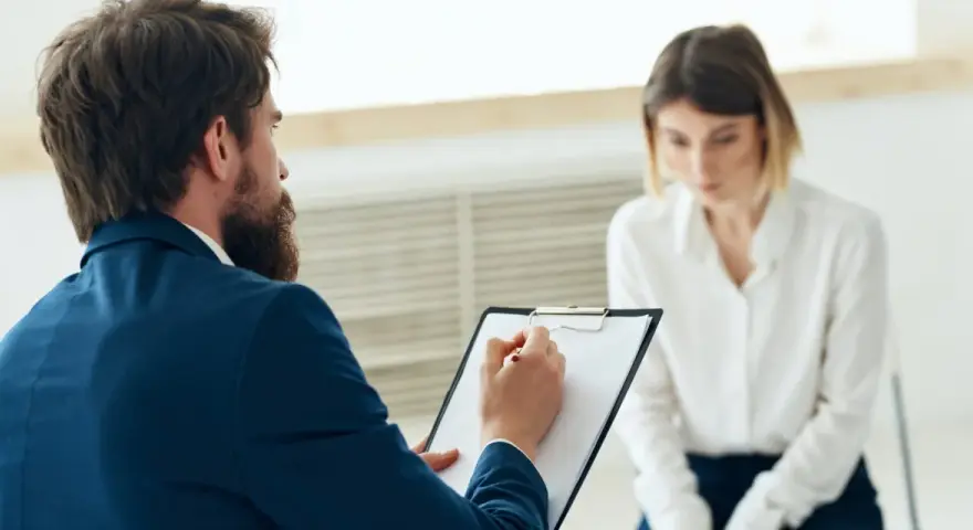 Mężczyzna z brodą w garniturze notuje coś na kartce, siedząc naprzeciwko kobiety.