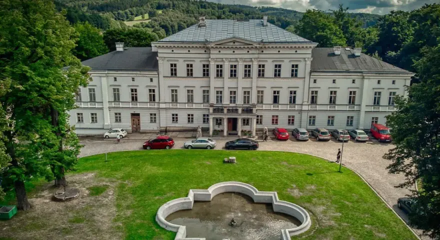 Pałac Jedlinka, biała, okazała budowla z szarym dachem, otoczona zielenią i górami. Przed pałacem fontanna i parking z samochodami.