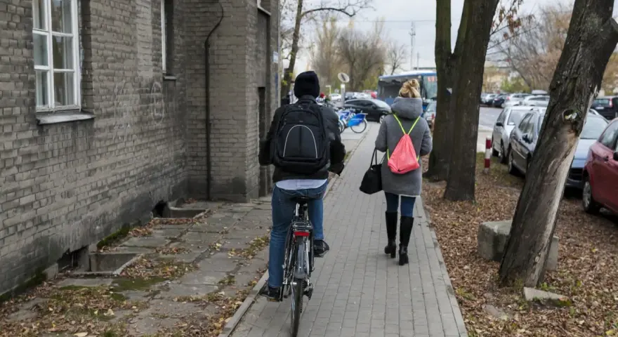 Osoba na rowerze i kobieta idąca chodnikiem obok budynku i drzew.