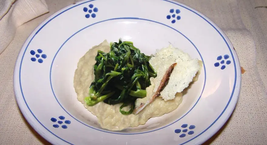 Piatto con fave e cicoria saltata, guarnito con una scaglia di formaggio e un'acciuga.