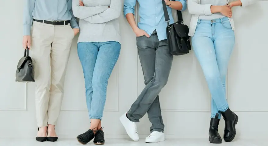 Cztery osoby w casual styl stoją przy ścianie. Jedna w jasnych spodniach i czarnych szpilkach, druga w jeansach i czarnych botkach, trzecia w szarych jeansach i białych sneakersach, czwarta w jasnych jeansach i czarnych półbutach.