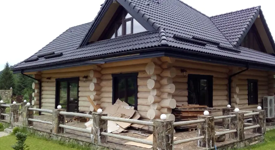 Drewniany dom z bali z czarnym dachem i tarasem, otoczony zielenią.