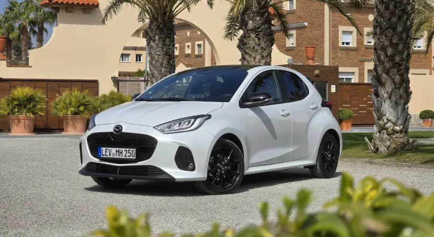 Biała Mazda 2 Hybrid zaparkowana przed budynkiem z palmami. Stylowy miejski samochód gotowy na podróż.