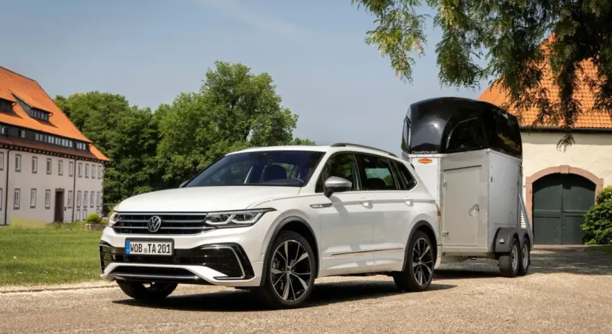 Biały Volkswagen SUV z przyczepą do przewozu koni stoi przed starym budynkiem.