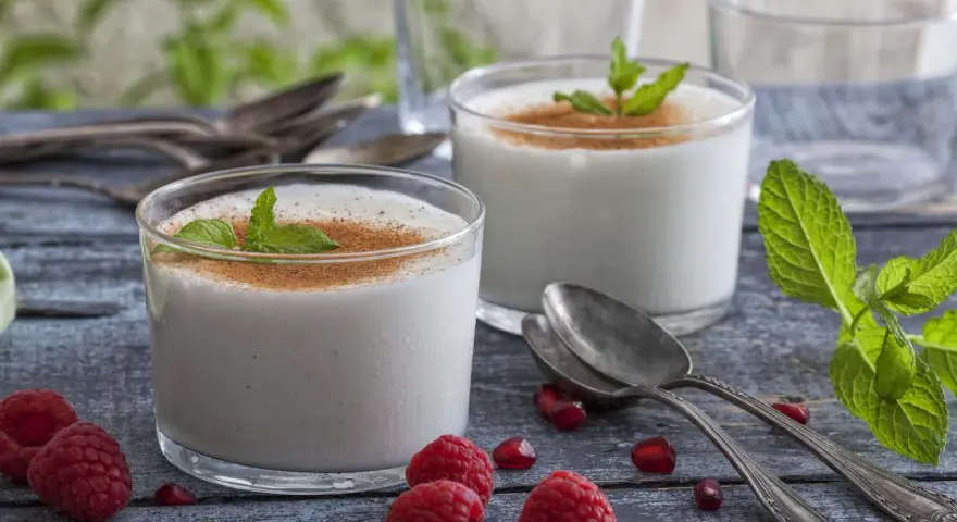 Zwei Gläser cremiger Leche Merengada, garniert mit Minze und Zimt, umgeben von Himbeeren und Granatapfelkernen.