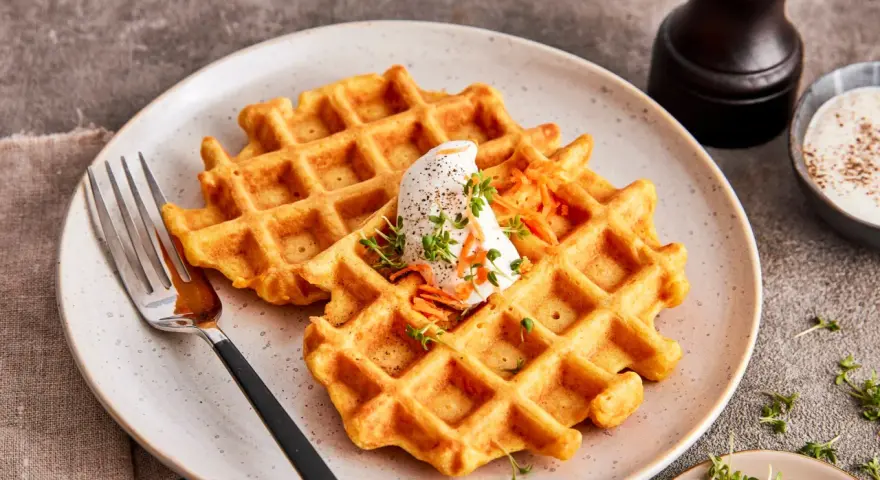 Zwei goldbraune Karotten Waffeln mit einem Klecks Sahne und geraspelten Karotten, garniert mit Kresse.