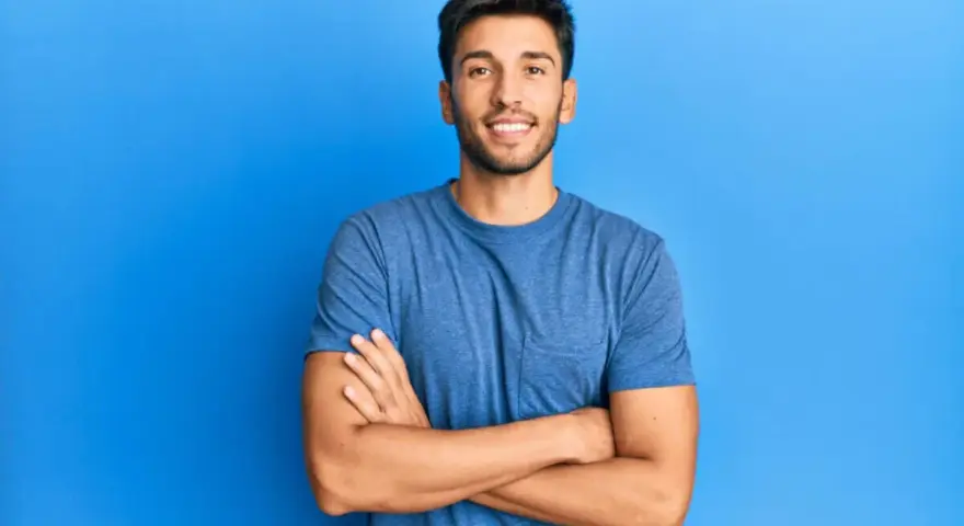 Hombre sonriente con estilos barba poco poblada, brazos cruzados, sobre fondo azul.