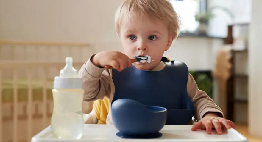 Mały chłopiec w śliniaku próbuje samodzielnie jeść. Obok butelka i miseczka z serii naczyń silikonowych dla dzieci.