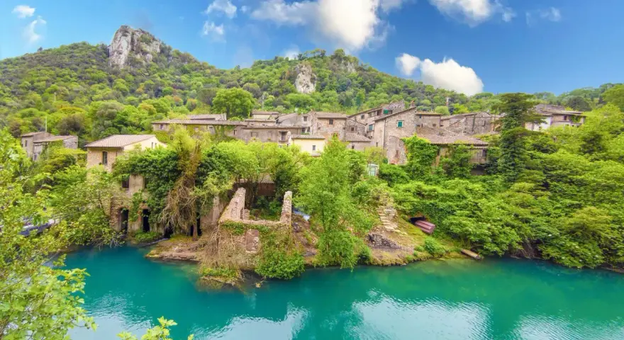 Un borgo incantevole in Umbria, con case in pietra ricoperte di vegetazione che si affacciano su un fiume turchese. Un luogo perfetto per i cammini.