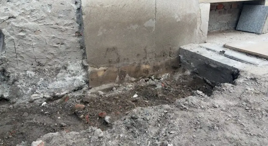 Widoczne ślady osiadania budynku i pęknięcia na cokole. Uszkodzony fragment fundamentu, ziemia i gruz wokół.